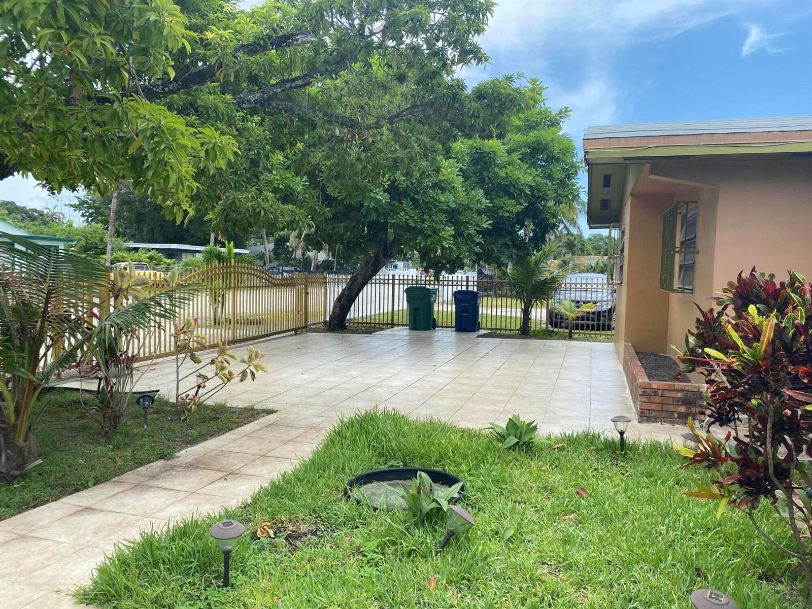 A paved patio area sits beside a house, featuring a large tree and metal fencing — 1050 NW 100th Street, Miami FL 33150, MIAMI, FL