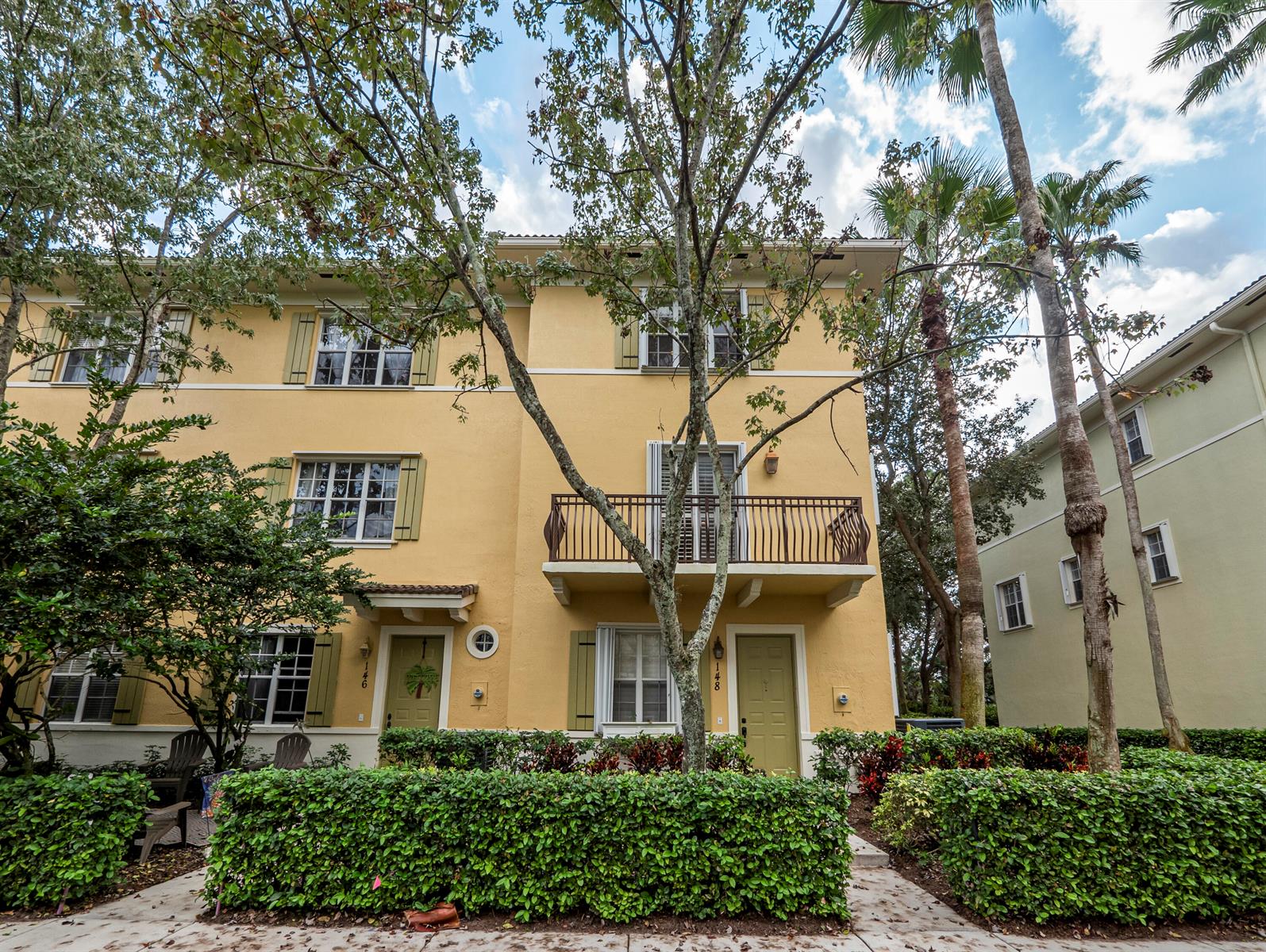 A three-story yellow townhouse with a balcony, green shutters, and a manicured hedge in front — 148 E Morning Dew Circle, Jupiter FL 33458, JUPITER, FL