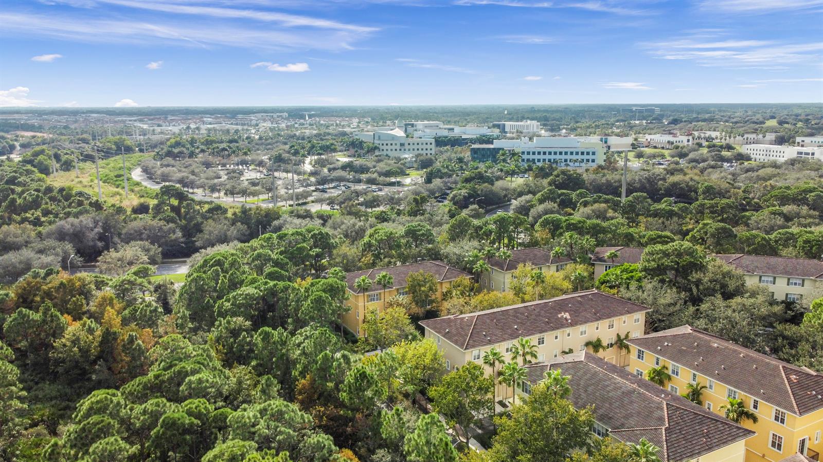 An aerial view shows multi-story apartment buildings surrounded by dense trees and a distant office park — 148 E Morning Dew Circle, Jupiter FL 33458, JUPITER, FL