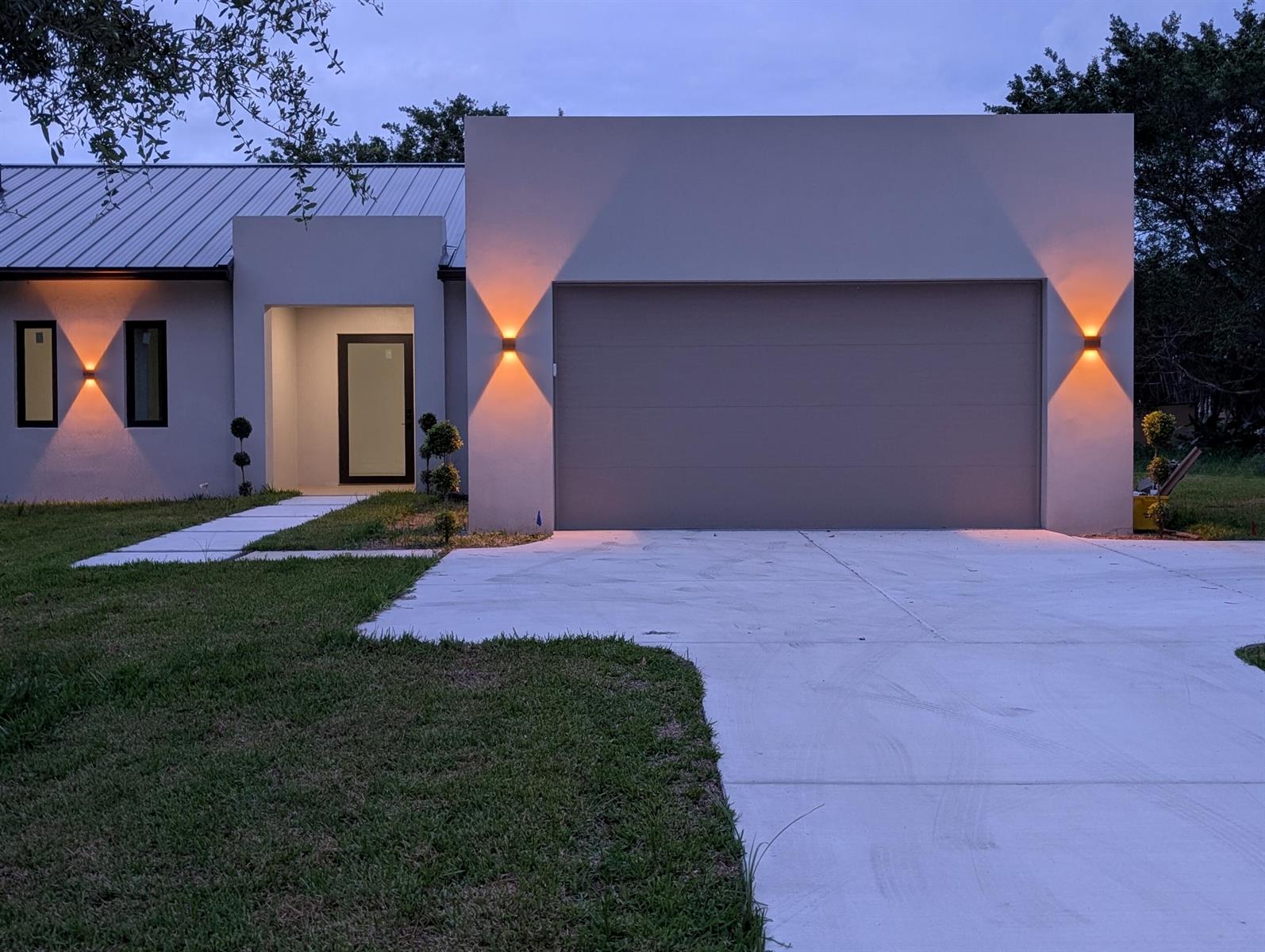 A modern house exterior features a flat-roofed garage, a recessed entryway, and illuminated wall sconces — 5356 Colbright Road, Lake Worth FL 33467, LAKE WORTH, FL