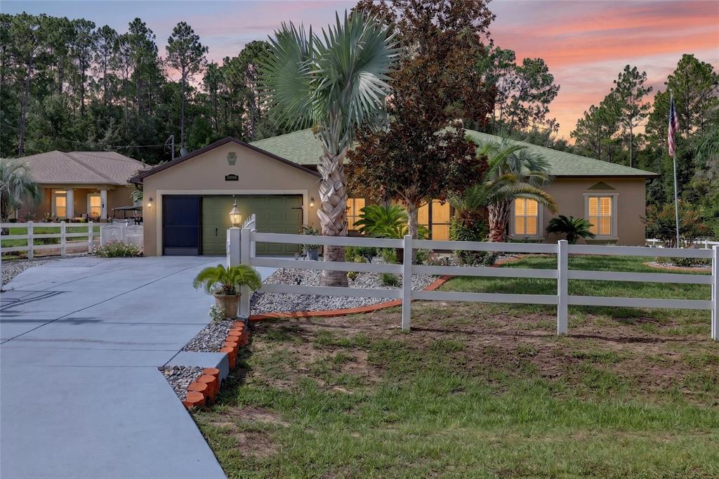 A single-story house with a green garage door, white fence, and palm trees at sunset — 20096 SW 59TH LANE, DUNNELLON, FL