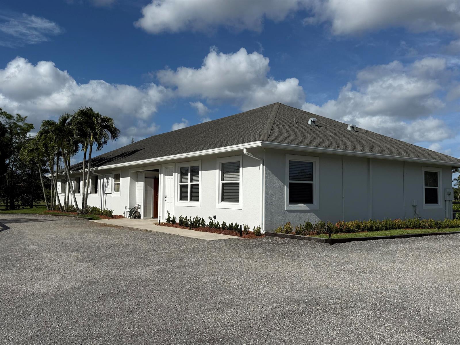 A white single-story building with a gray shingled roof sits beside a gravel driveway — 6600 Duckweed Road, Wellington FL 33449, WELLINGTON, FL
