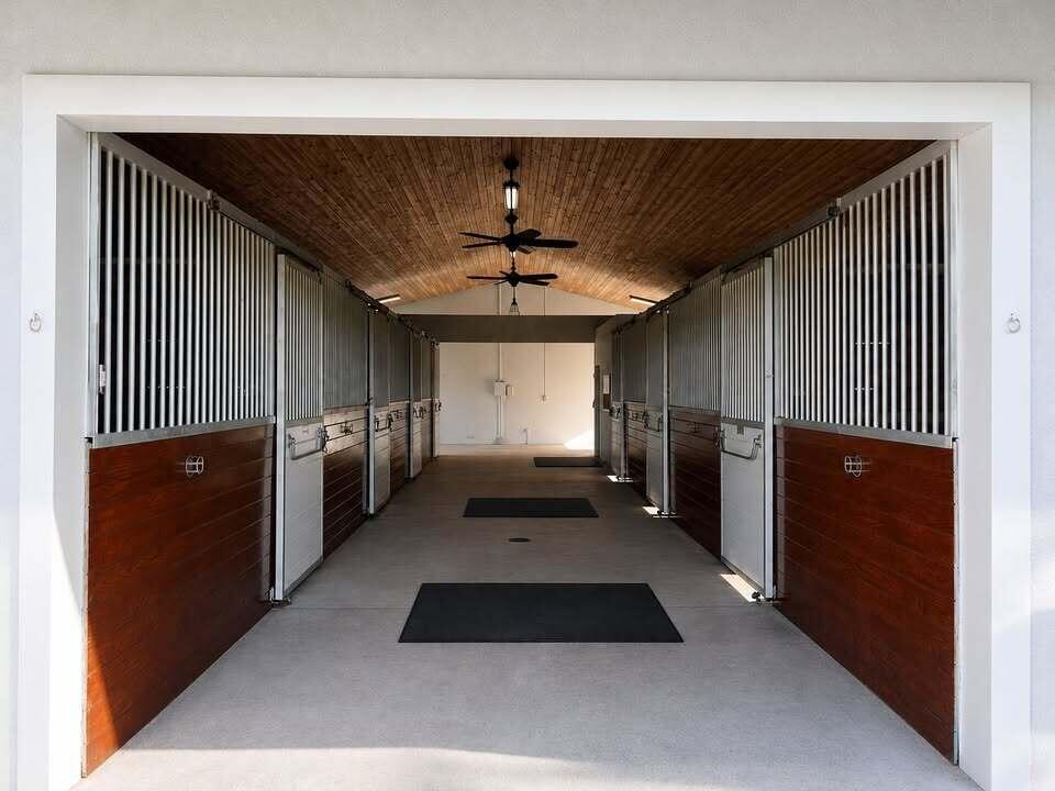 A horse stable aisle featuring wood-paneled stalls with metal bars and a vaulted wood ceiling — 6600 Duckweed Road, Wellington FL 33449, WELLINGTON, FL