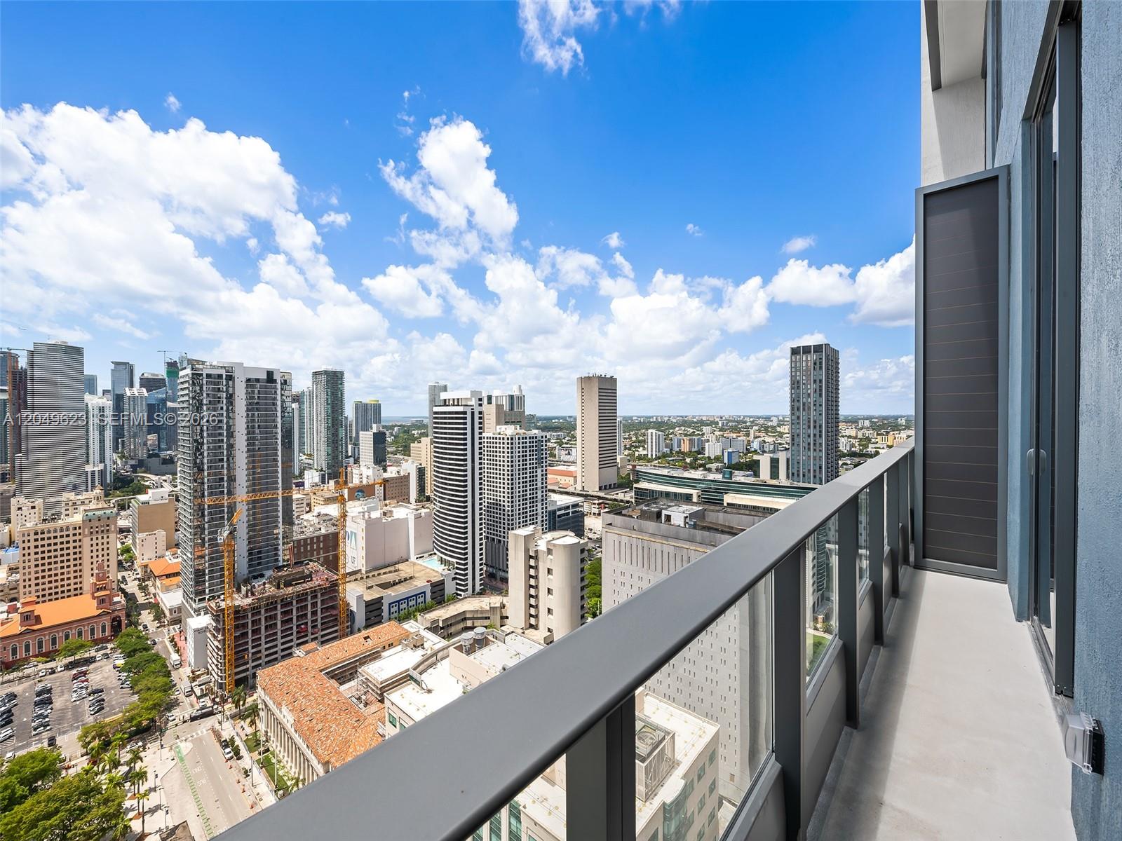 A high-rise balcony with a glass railing overlooks a dense urban cityscape under a blue sky — 121 NE 5th St # 3703, Miami FL 33132, MIAMI, FL
