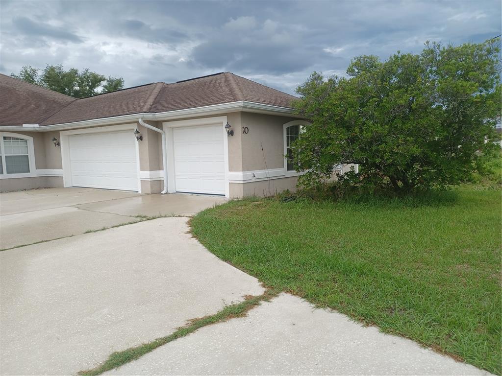 A tan duplex with two white garage doors and a concrete driveway under a cloudy sky — 10 ROCKEFELLER DRIVE, PALM COAST, FL