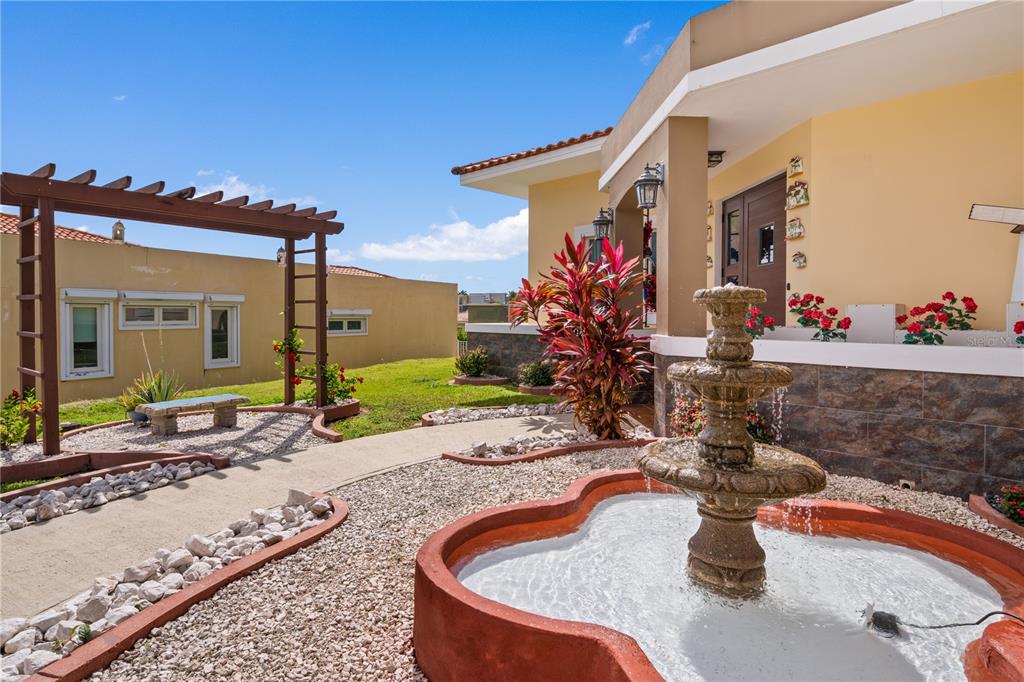 A tiered stone fountain sits in a gravel courtyard beside a house with a pergola — Palmas del Mar THE VIEWS # 12, HUMACAO, PR