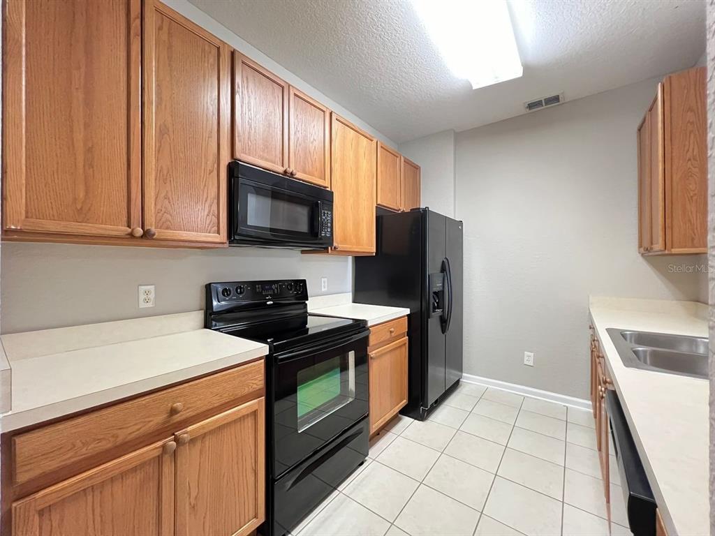 A kitchen features oak cabinets, black appliances, light countertops, and beige tile flooring — 2484 SAN TECLA STREET UNIT 204, ORLANDO, FL