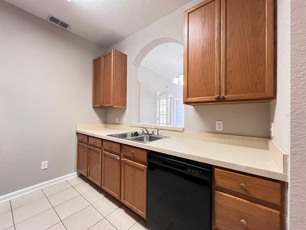 A kitchen features oak cabinets, a black dishwasher, a double sink, and beige tile flooring — 2484 SAN TECLA STREET UNIT 204, ORLANDO, FL