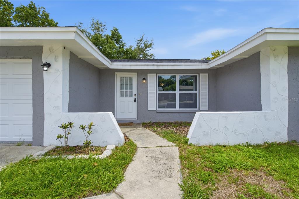 A gray house exterior features a white front door, a window with shutters, and landscaping — 1811 FOREST DRIVE, OLDSMAR, FL