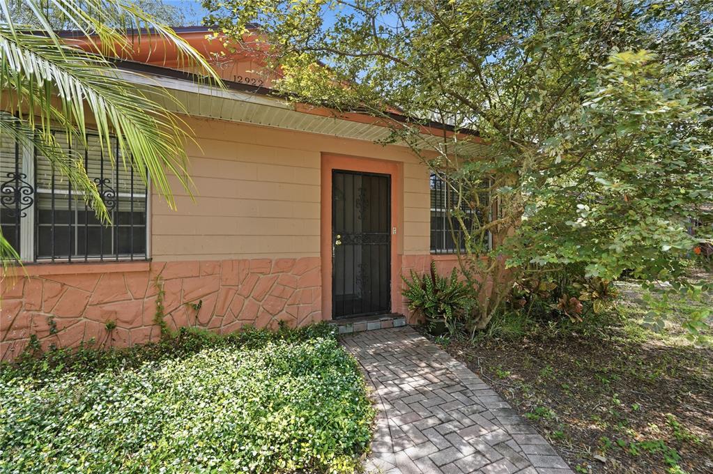 A single-story house exterior features a brick walkway leading to a dark metal front door — 12922 N ALBANY AVENUE, TAMPA, FL