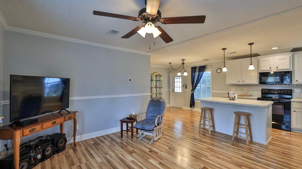 A living room with light blue walls and wood flooring opens into a kitchen with white cabinets — 1157 MILLBROOK AVENUE, PORT ORANGE, FL