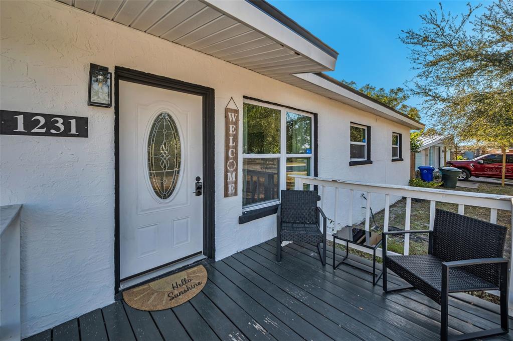 A white house exterior features a dark wooden porch with two chairs and a door — 1231 LITTLE JOHN LANE, DUNEDIN, FL