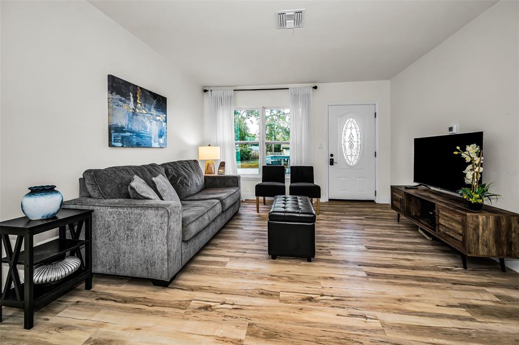 A living room features a gray sofa, a wood media console, and wood-look flooring — 1231 LITTLE JOHN LANE, DUNEDIN, FL