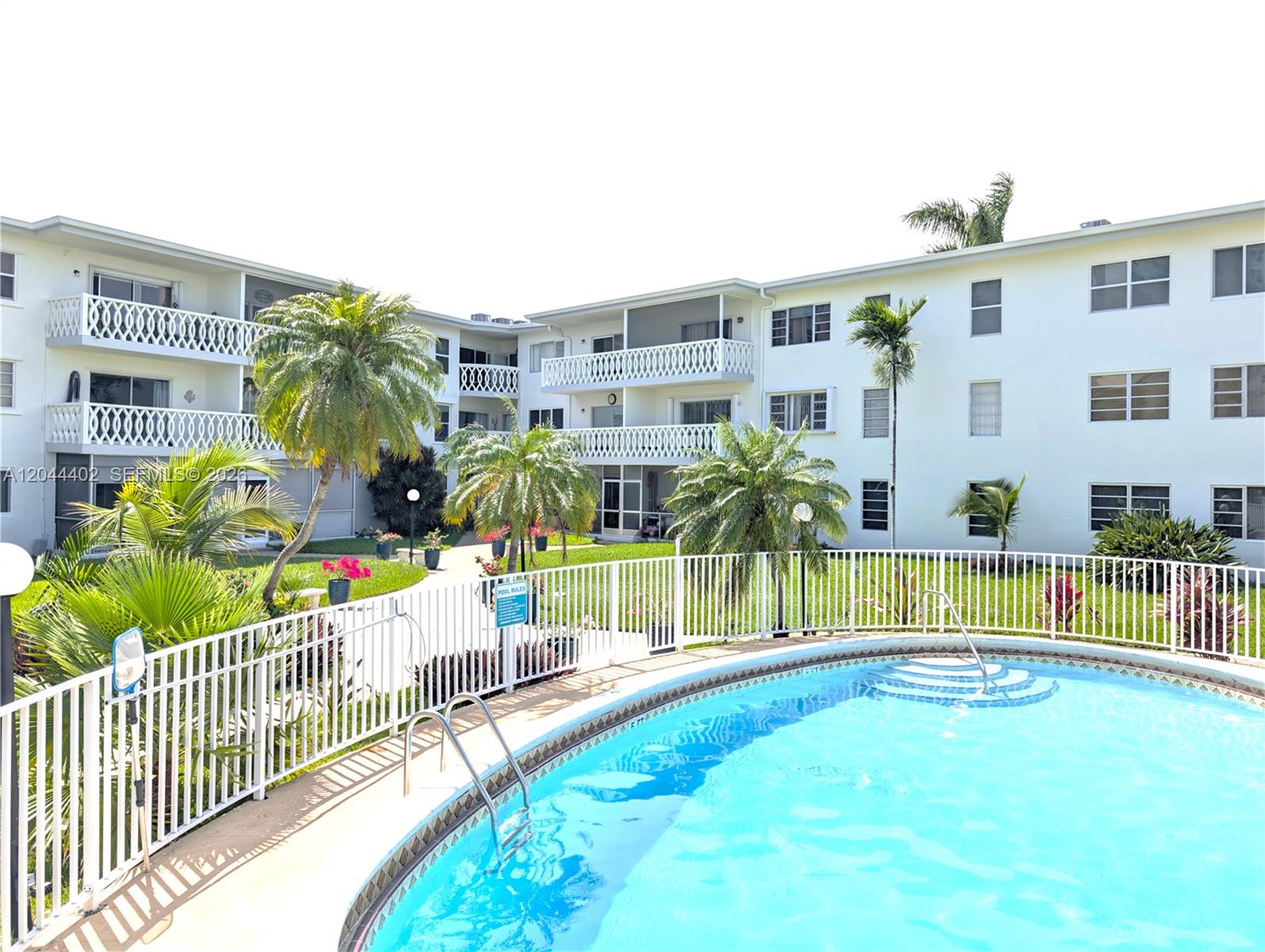 An outdoor swimming pool is surrounded by a white fence and multi-story apartment buildings — 450 GOLDEN ISLES DR # 2H, Hallandale Beach FL 33009, HALLANDALE BEACH, FL