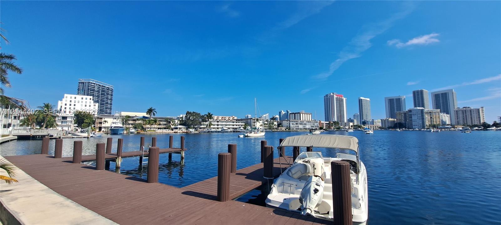 A boat is docked at a wooden pier overlooking a waterway with city buildings beyond — 450 GOLDEN ISLES DR # 2H, Hallandale Beach FL 33009, HALLANDALE BEACH, FL