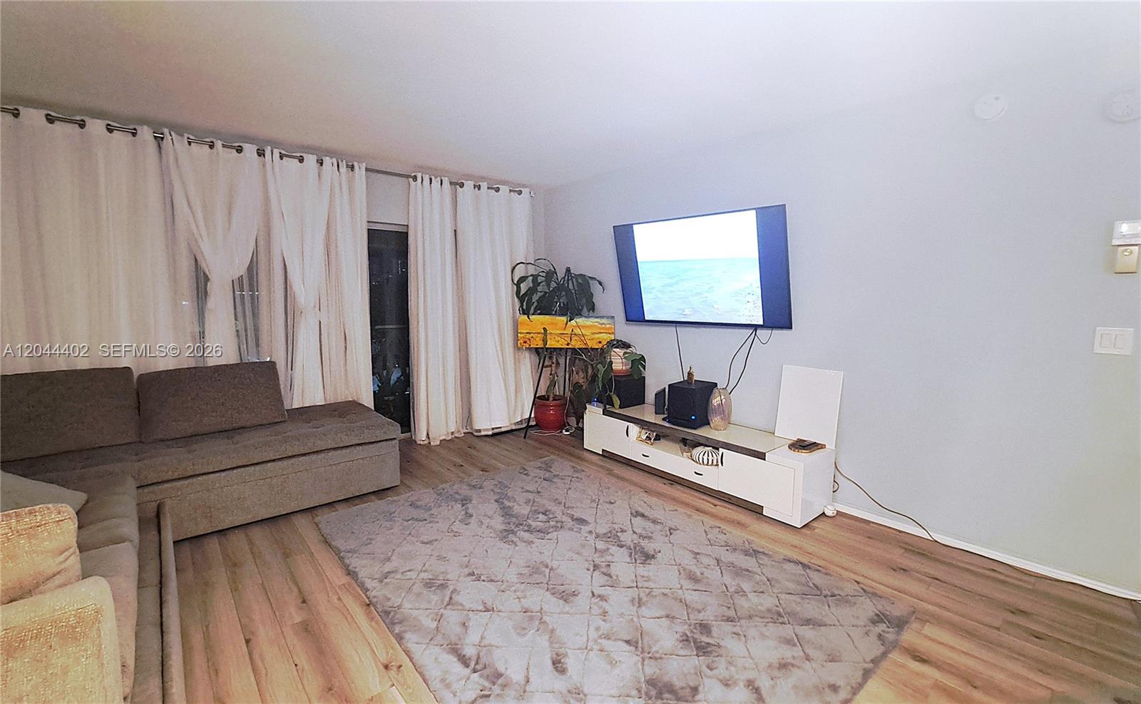 A living room features light wood flooring, a grey sectional sofa, and a wall-mounted television — 450 GOLDEN ISLES DR # 2H, Hallandale Beach FL 33009, HALLANDALE BEACH, FL