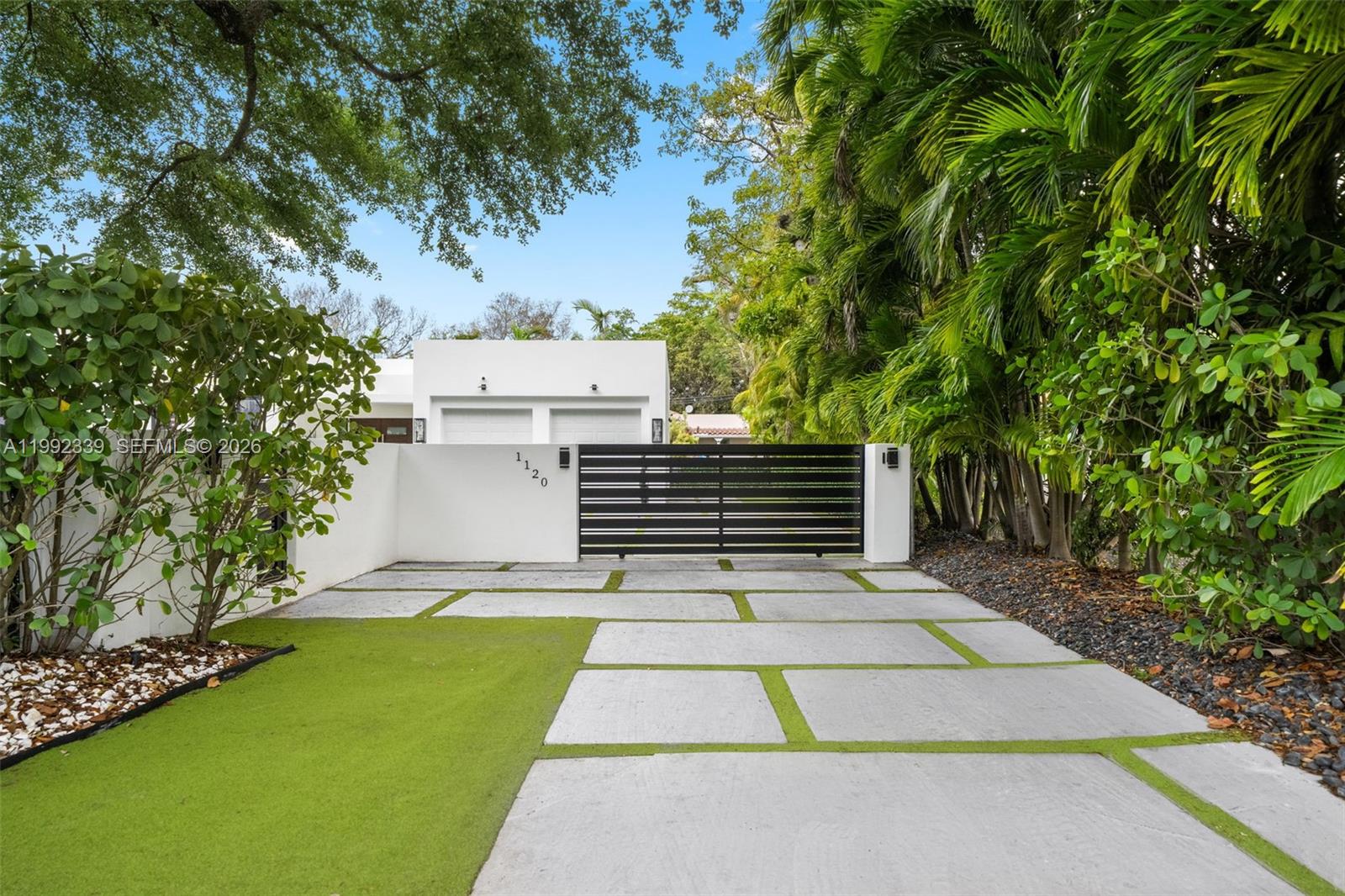 A modern white home features a black horizontal gate, concrete pavers, and lush landscaping — 1120 Wallace St, Coral Gables FL 33134, CORAL GABLES, FL