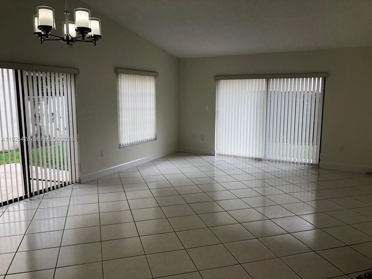 An empty room with light-colored tile flooring, a chandelier, and vertical blinds on sliding doors — 9044 SW 112 CT, Miami FL 33176, MIAMI, FL