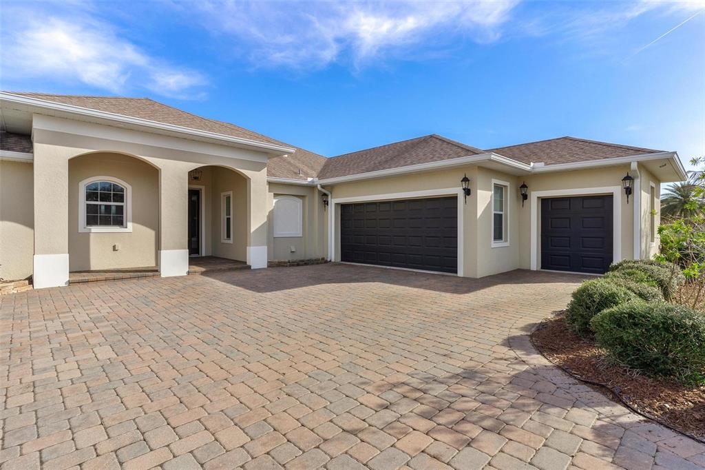 A beige house exterior features a paver driveway, two dark garage doors, and arched entry — 39634 GROVE HEIGHTS, LADY LAKE, FL