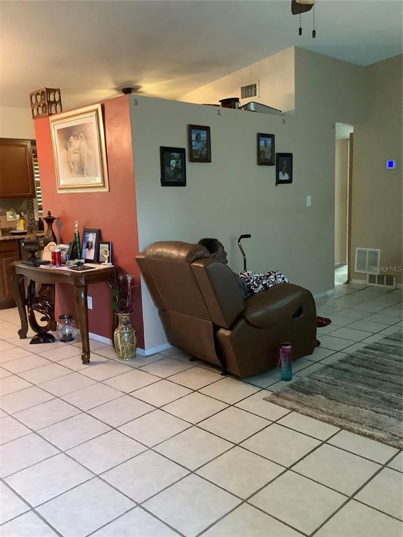 A person sits in a brown recliner in a tiled living room with wall-mounted photos — 819 62ND AVENUE S, ST PETERSBURG, FL