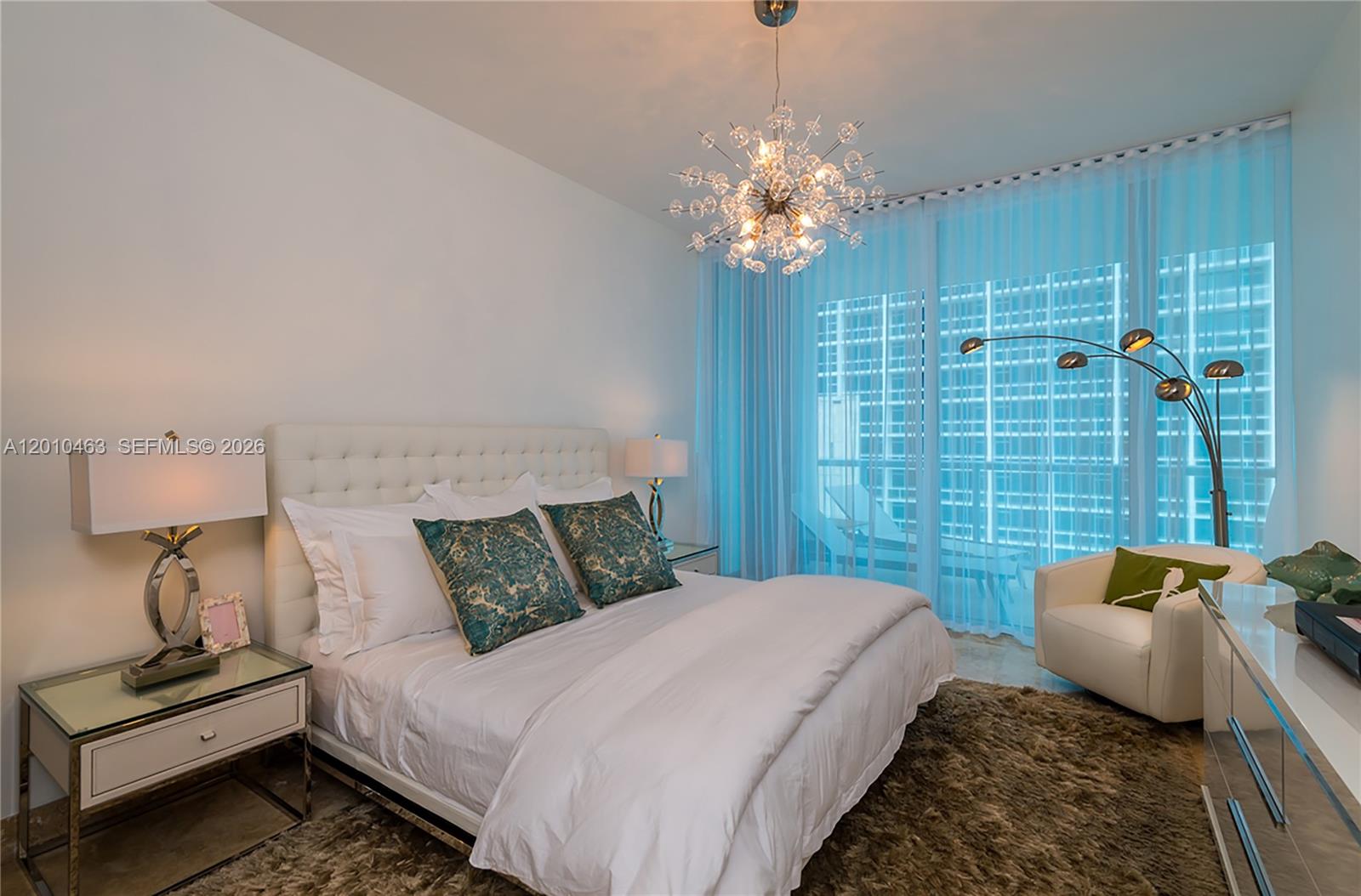 A bedroom features a tufted headboard, a sputnik chandelier, and floor-to-ceiling windows with sheer curtains — 6799 Collins Ave # 503, Miami Beach FL 33141, MIAMI BEACH, FL
