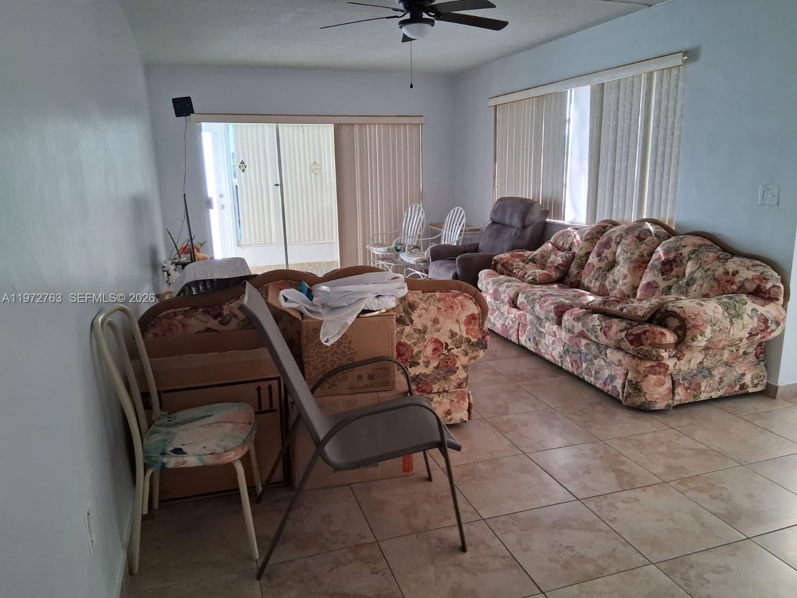 A living room features floral sofas, tile flooring, a ceiling fan, and sliding glass doors — 300 NE 191st St # 211, Miami FL 33179, MIAMI, FL