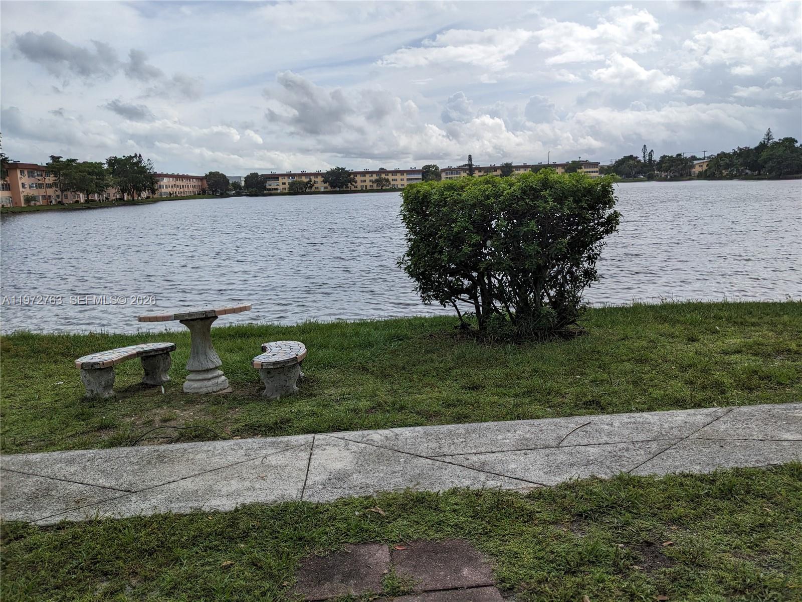 A stone table and benches sit on a grassy lawn overlooking a lake — 300 NE 191st St # 211, Miami FL 33179, MIAMI, FL