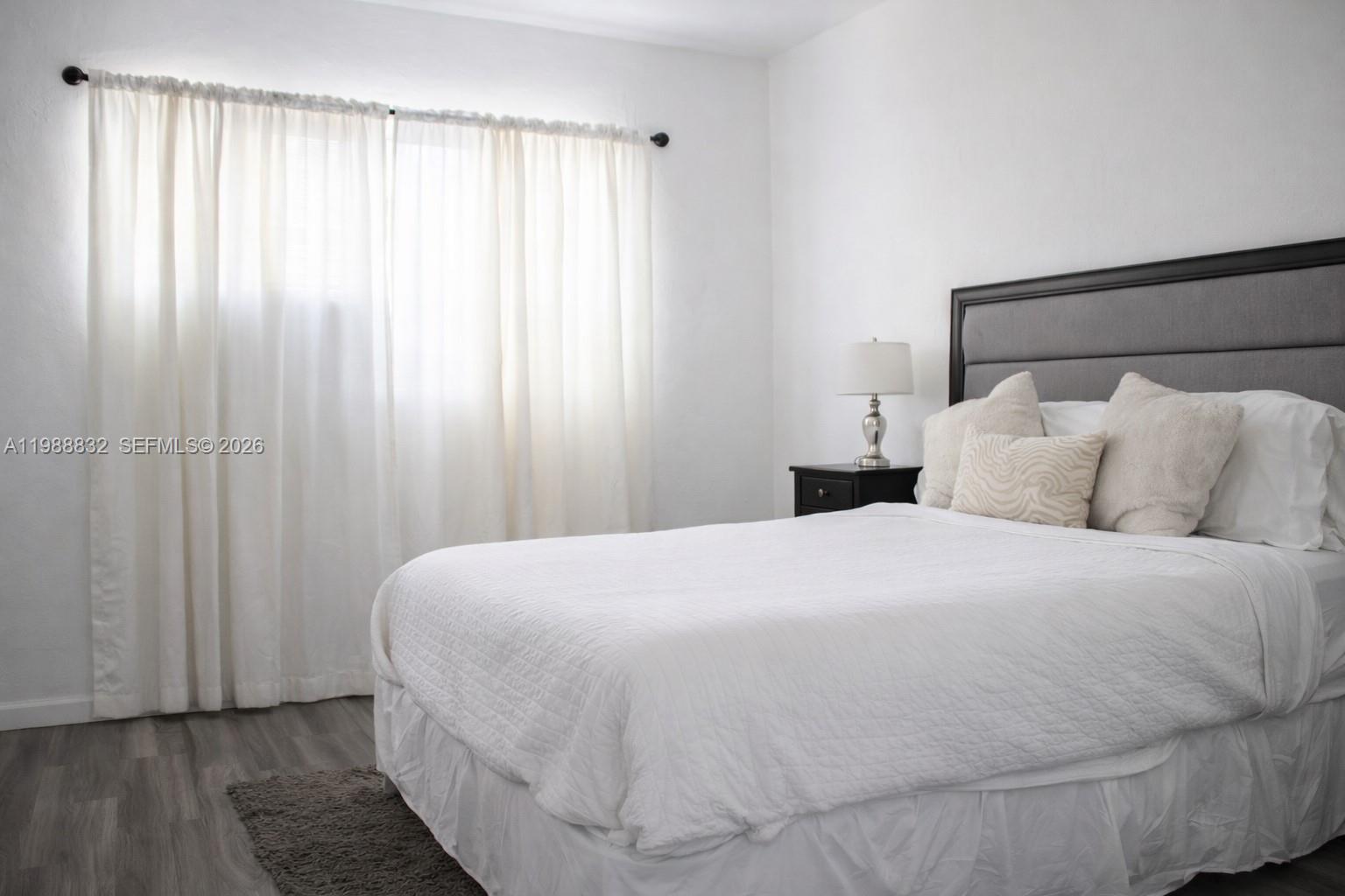 A bedroom features a bed with a gray headboard, white bedding, and sheer window curtains — 6250 NW Miami Pl # 2, Miami FL 33150, MIAMI, FL