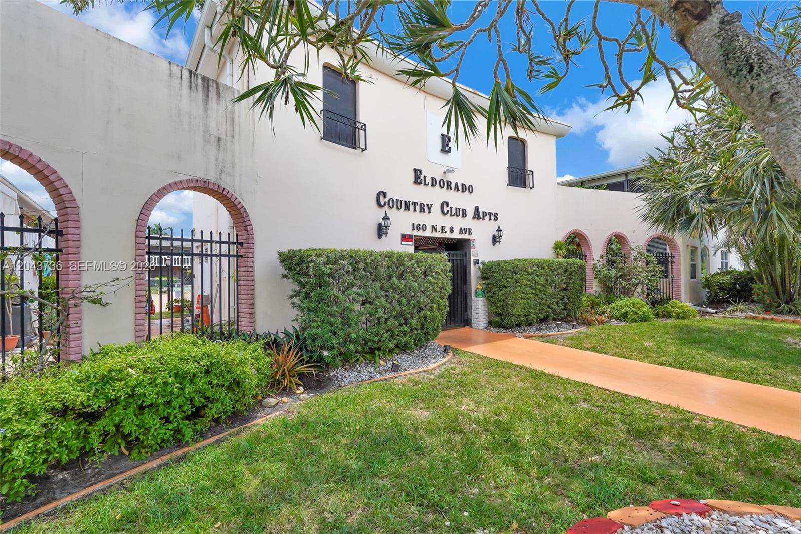 The exterior of a two-story apartment building features arched brick entryways and landscaped grounds — 160 NE 8th Ave # 27A, Hallandale Beach FL 33009, HALLANDALE BEACH, FL