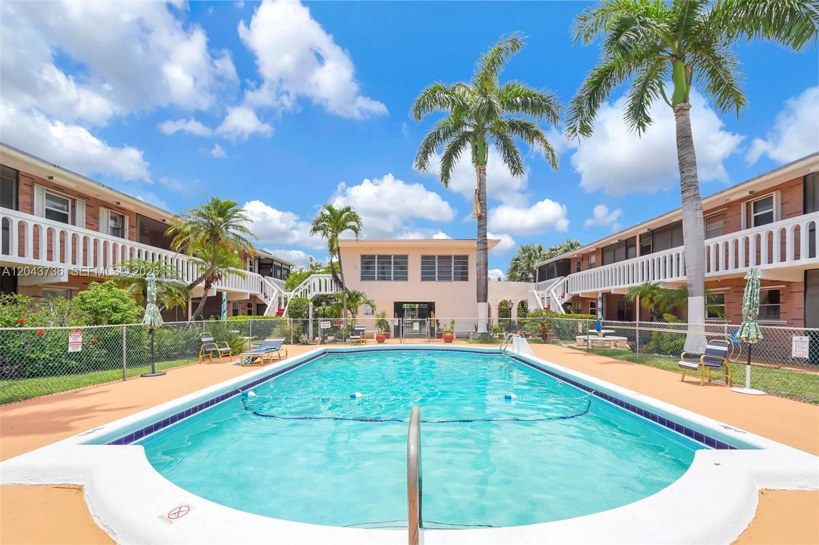An outdoor swimming pool is centered between two apartment buildings with palm trees and fencing — 160 NE 8th Ave # 27A, Hallandale Beach FL 33009, HALLANDALE BEACH, FL