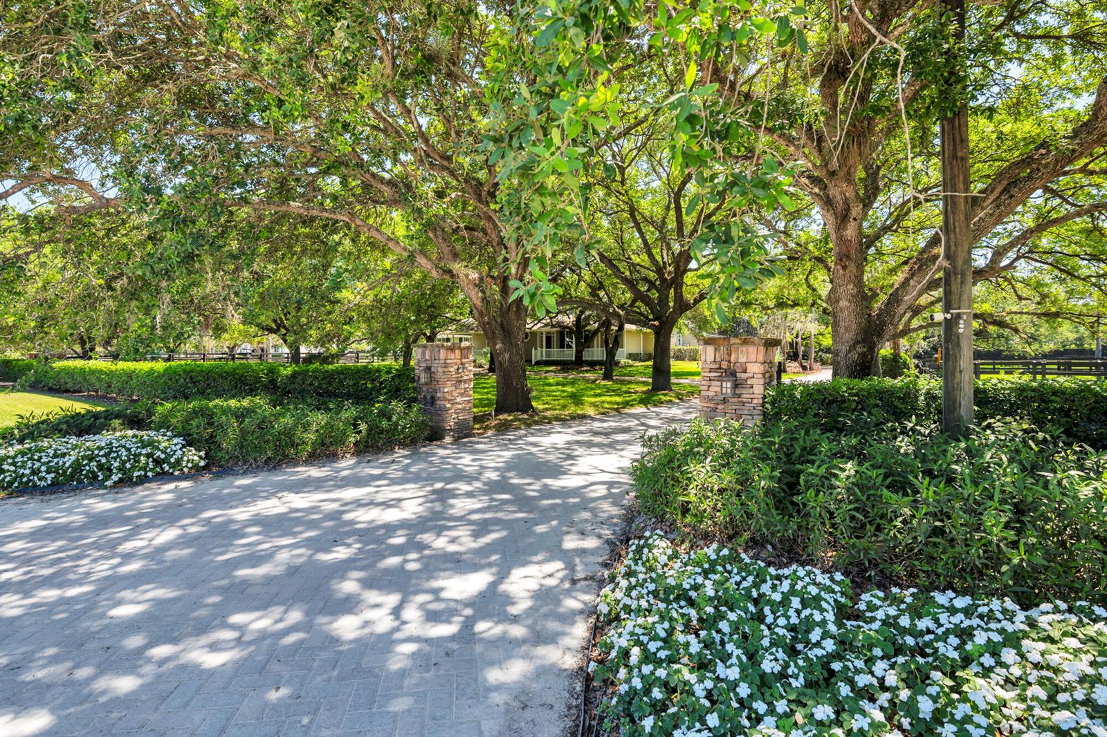 A paved driveway leads through stone pillars toward a house shaded by large oak trees — 14366 Belmont Trace, Wellington FL 33414, WELLINGTON, FL