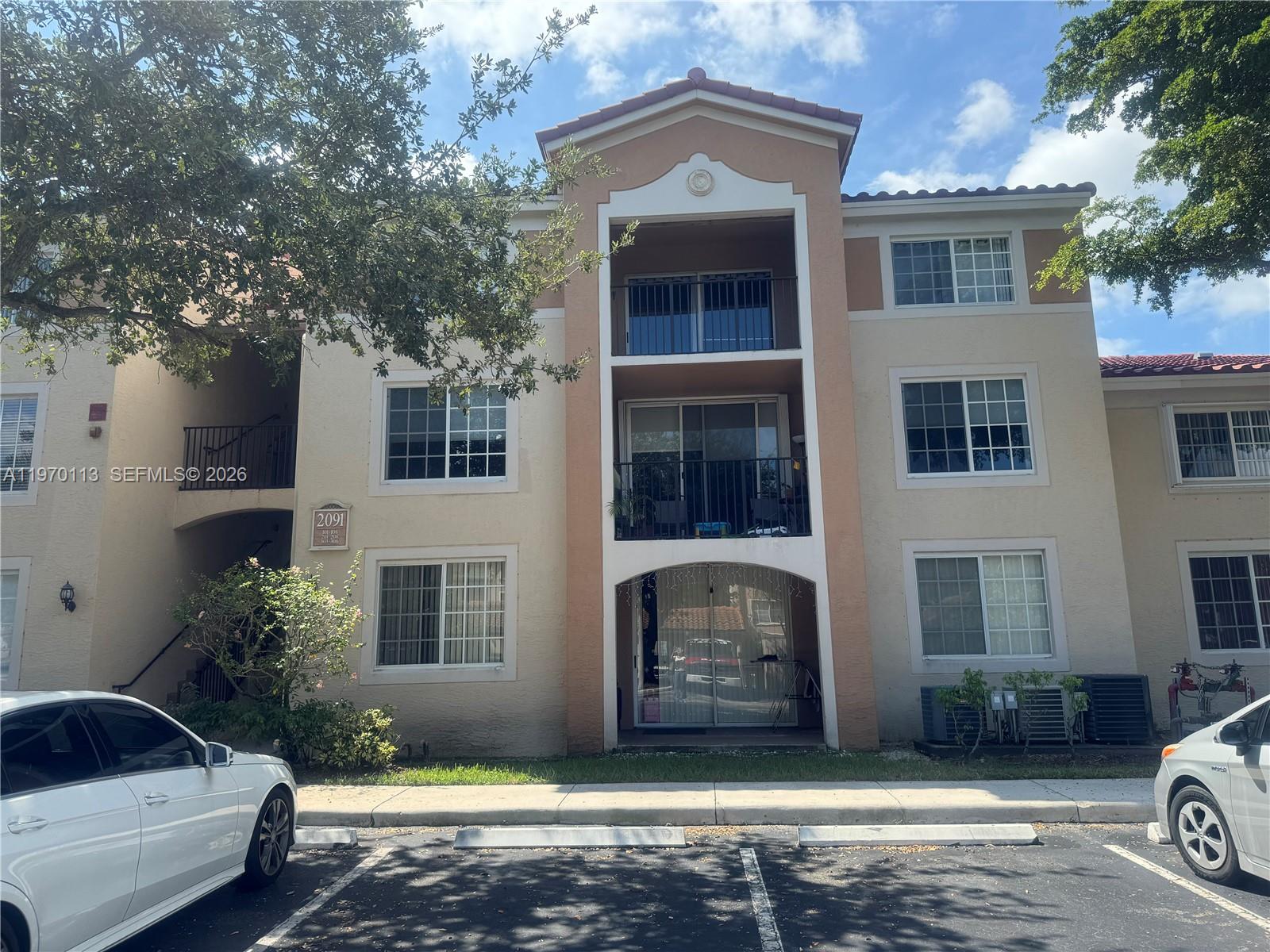 A three-story apartment building features beige stucco walls, balconies, and a parking lot in front — 2091 Renaissance Blvd # 104, Miramar FL 33025, MIRAMAR, FL