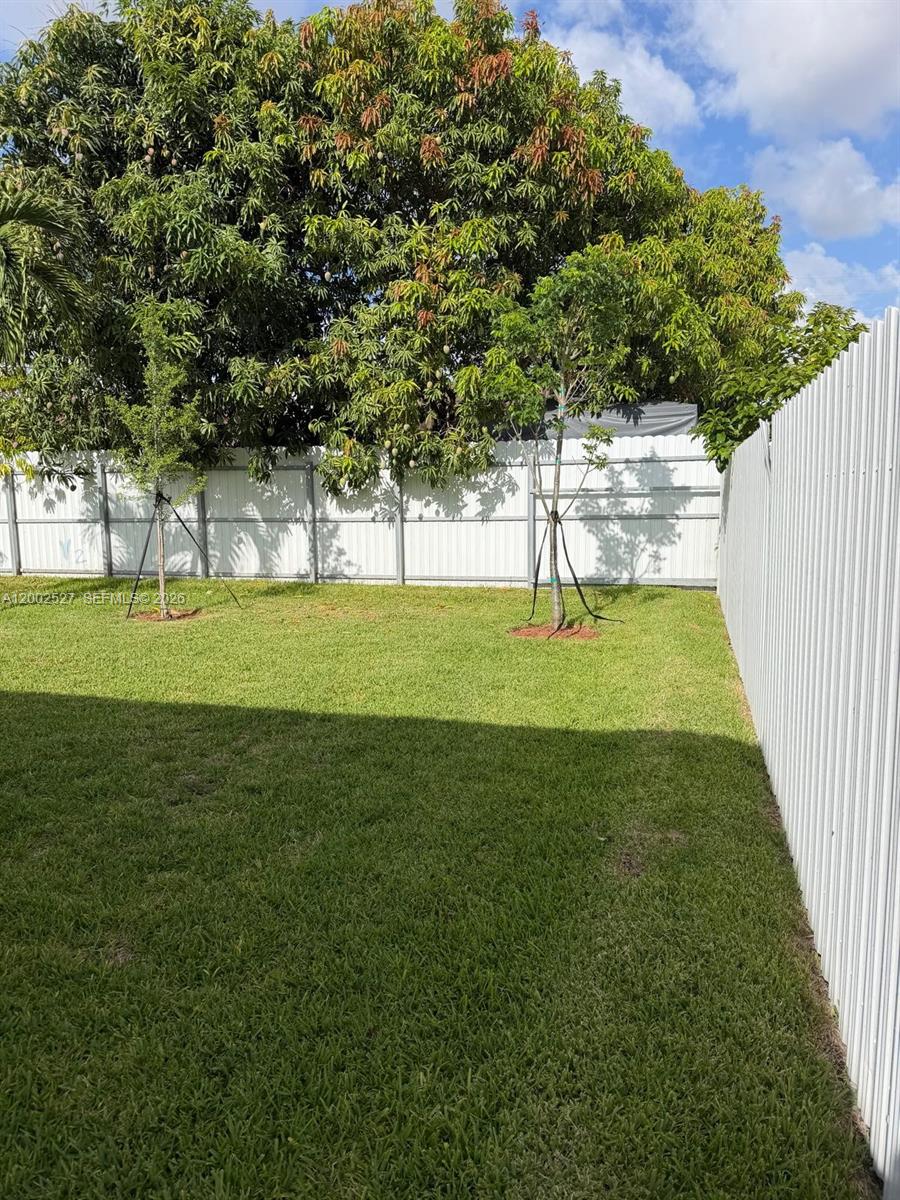A grassy backyard features two young trees and a white metal privacy fence — 831 SE 9th Ave, Hialeah FL 33010, HIALEAH, FL