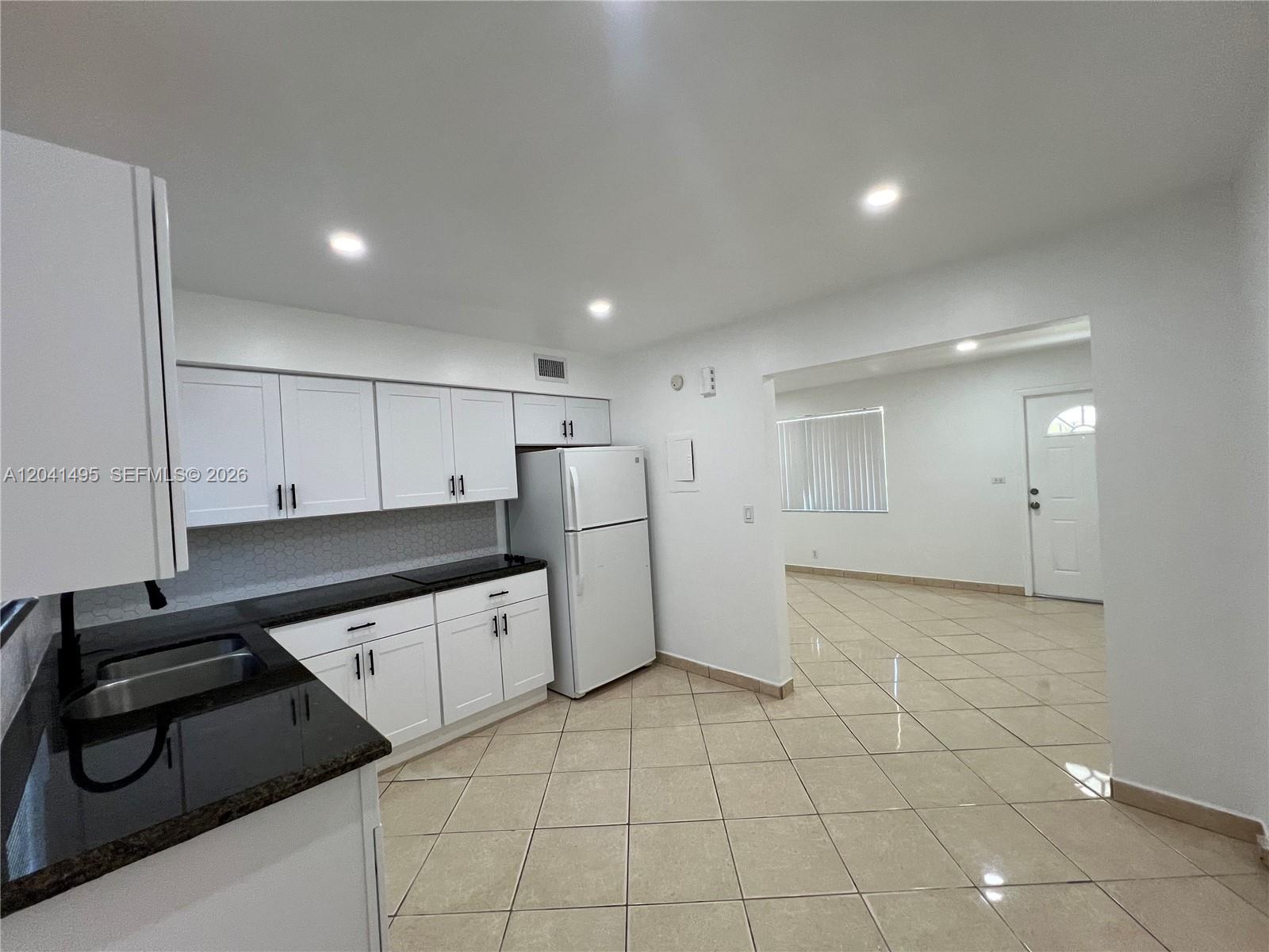 A kitchen with white cabinets, black countertops, and tile flooring opens into a living area — MIAMI, FL