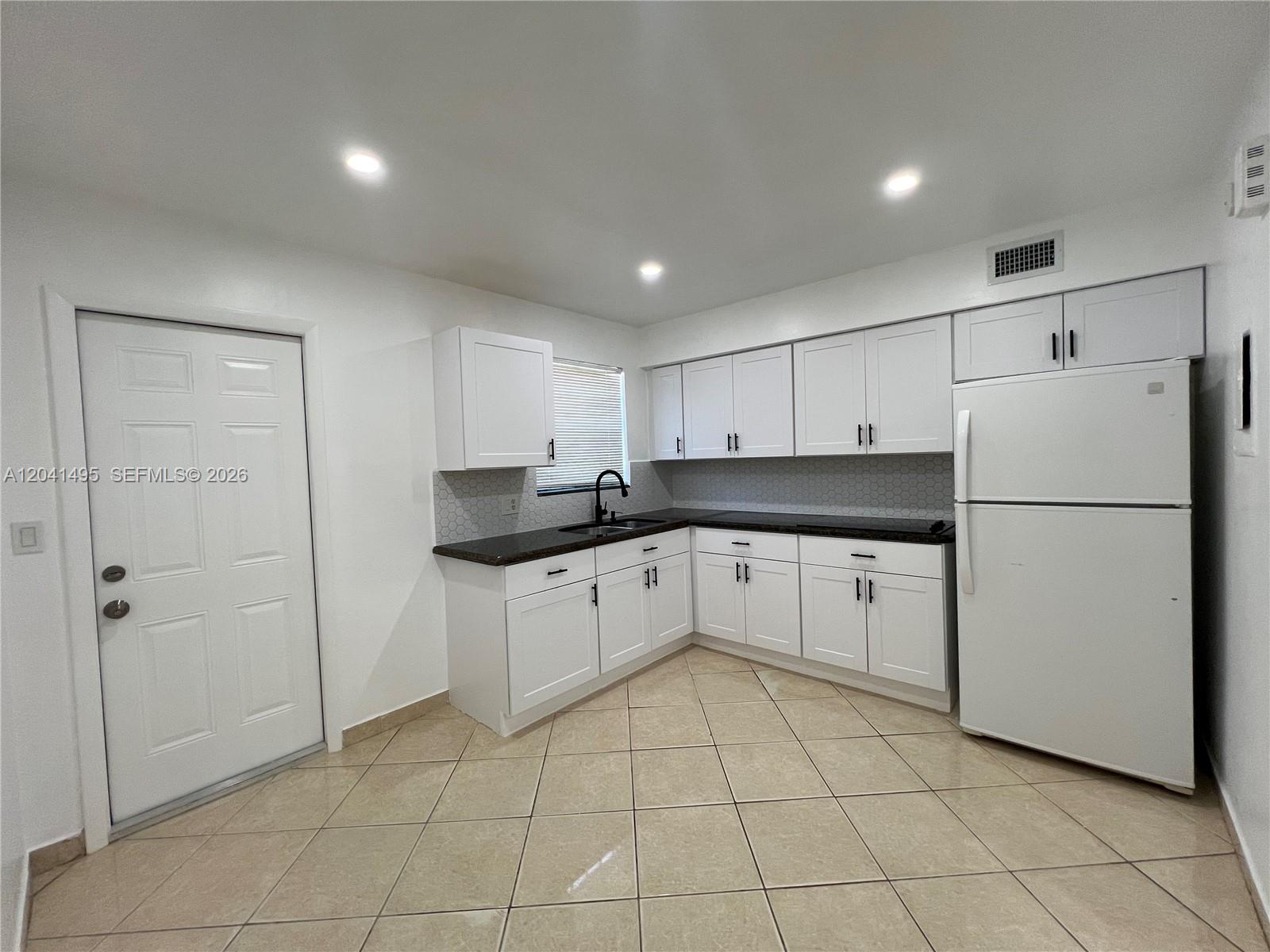A kitchen features white cabinets, dark countertops, a white refrigerator, and beige tile flooring — MIAMI, FL