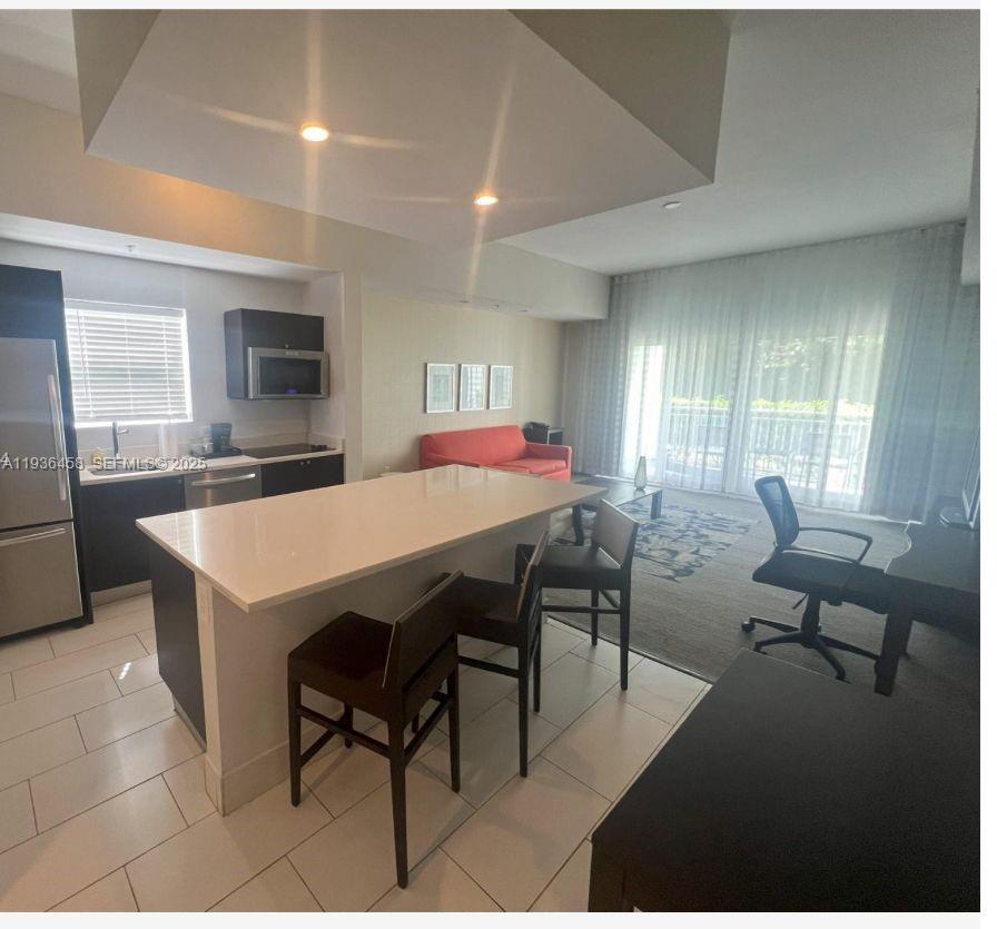 An open-concept kitchen with a white island and stools overlooks a living area with curtains — 5300 NW 87th Ave # 601, Doral FL 33178, DORAL, FL
