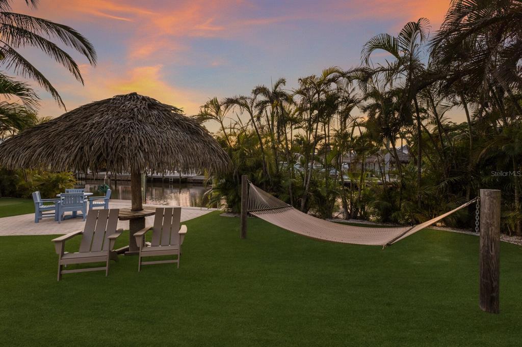 A backyard lawn features a tiki hut, patio furniture, and a hammock near a canal — 212 OAK AVENUE, ANNA MARIA, FL