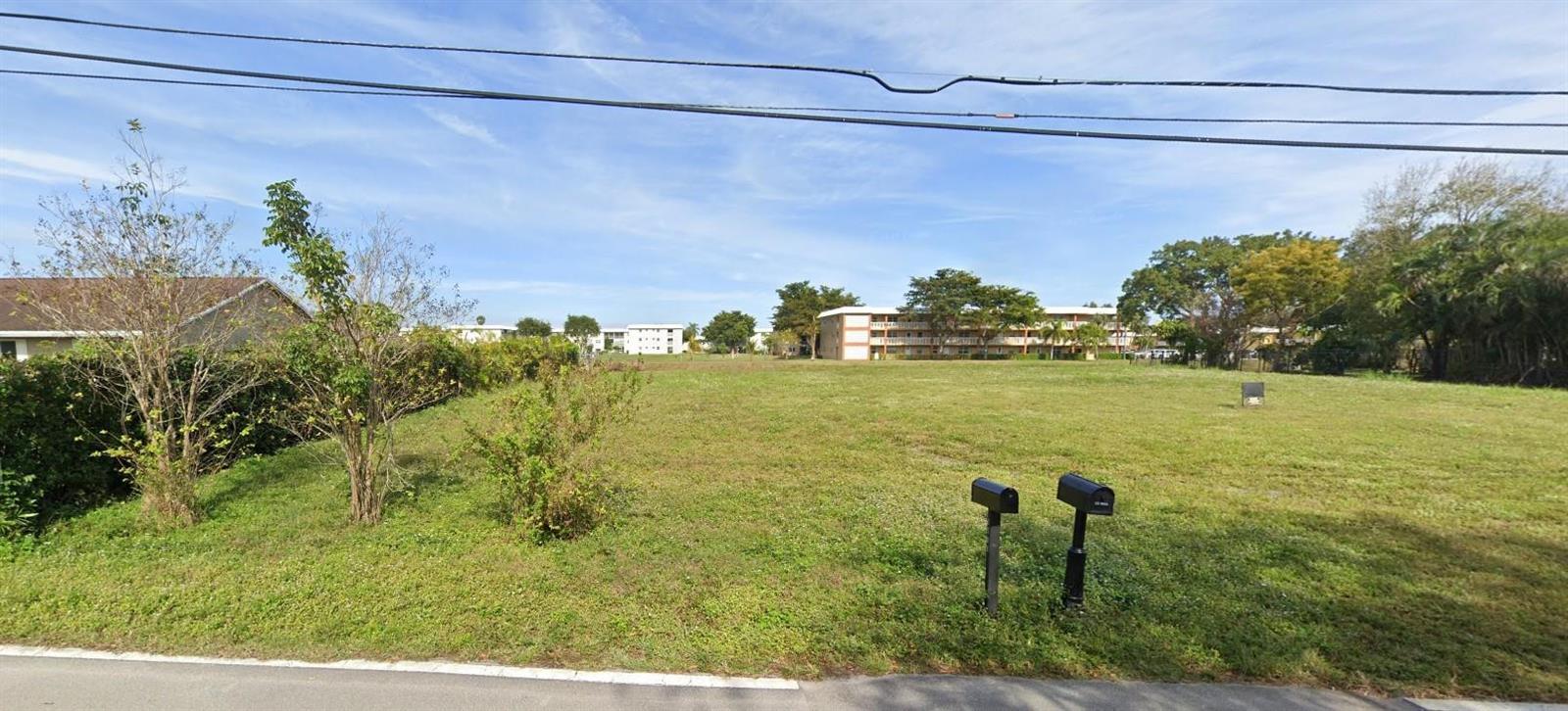 A grassy vacant lot sits between residential buildings under a clear blue sky — 5531 NW 77th Court, Pompano Beach FL 33073, POMPANO BEACH, FL