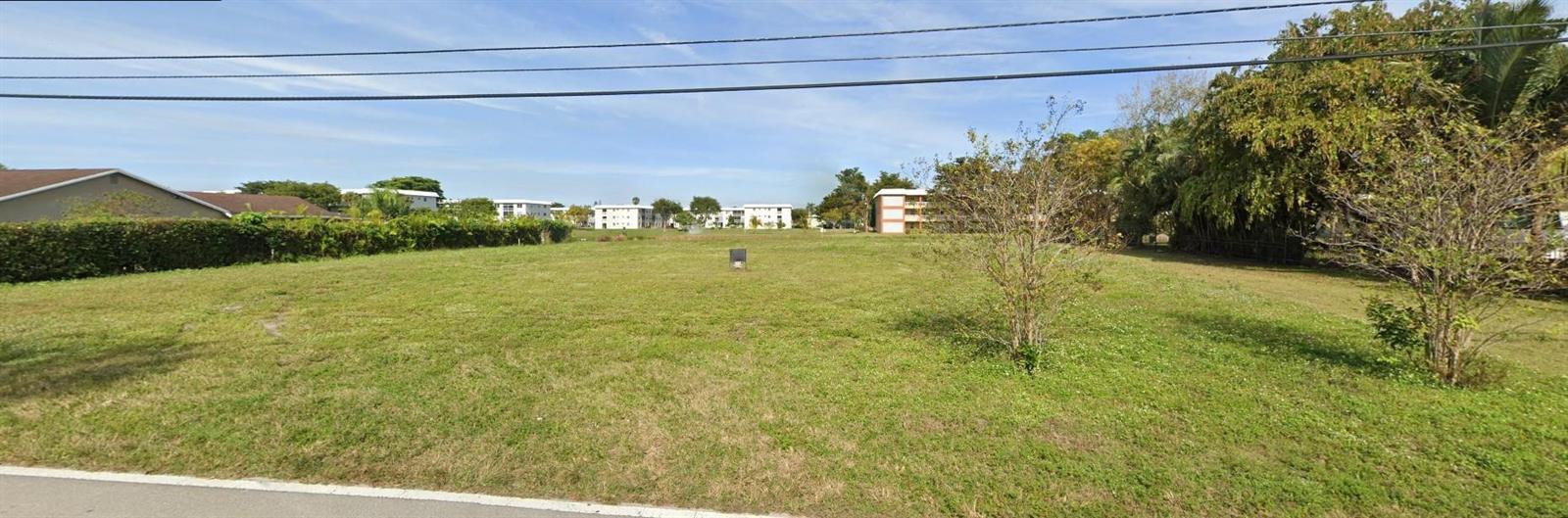 A vacant, grassy lot is bordered by trees and residential buildings under a blue sky — 5531 NW 77th Court, Pompano Beach FL 33073, POMPANO BEACH, FL