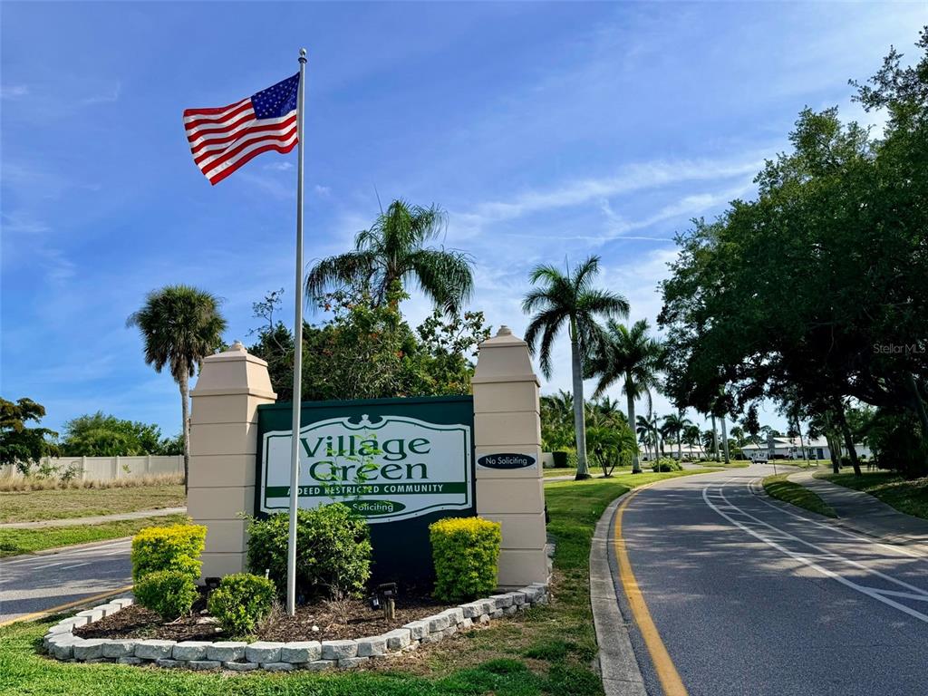 A sign for Village Green community stands beside a road with palm trees and flags — 7302 9TH AVENUE W UNIT 7302, BRADENTON, FL