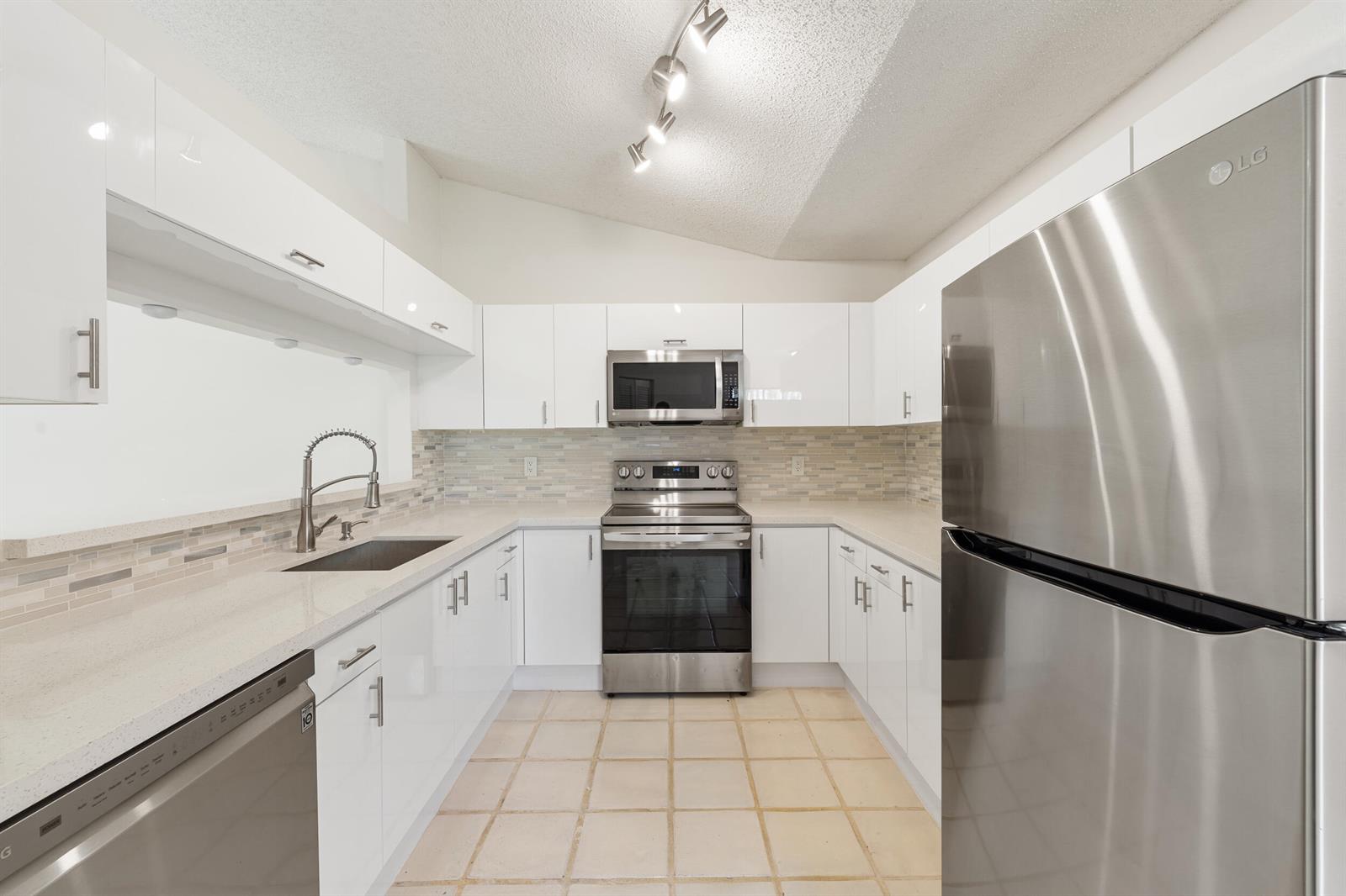 A kitchen features white glossy cabinets, stainless steel appliances, quartz countertops, and tiled flooring — 7219 Le Chalet Boulevard, Boynton Beach FL 33472, BOYNTON BEACH, FL