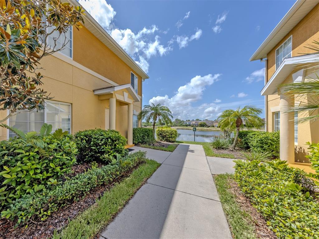 A sidewalk leads between two yellow residential buildings toward a small lake under blue skies — 12983 TIGERS EYE DRIVE, VENICE, FL