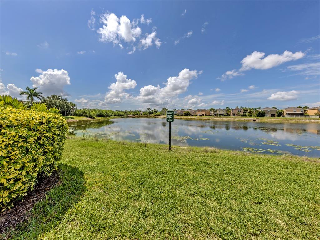 A grassy lawn leads to a pond with a warning sign and houses in the background — 12983 TIGERS EYE DRIVE, VENICE, FL