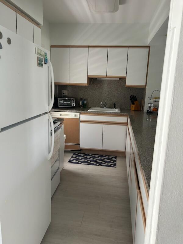 A galley kitchen features white cabinets with wood trim, a speckled countertop, and light flooring — 2760 Banyan Road # 25a, Boca Raton FL 33432, BOCA RATON, FL