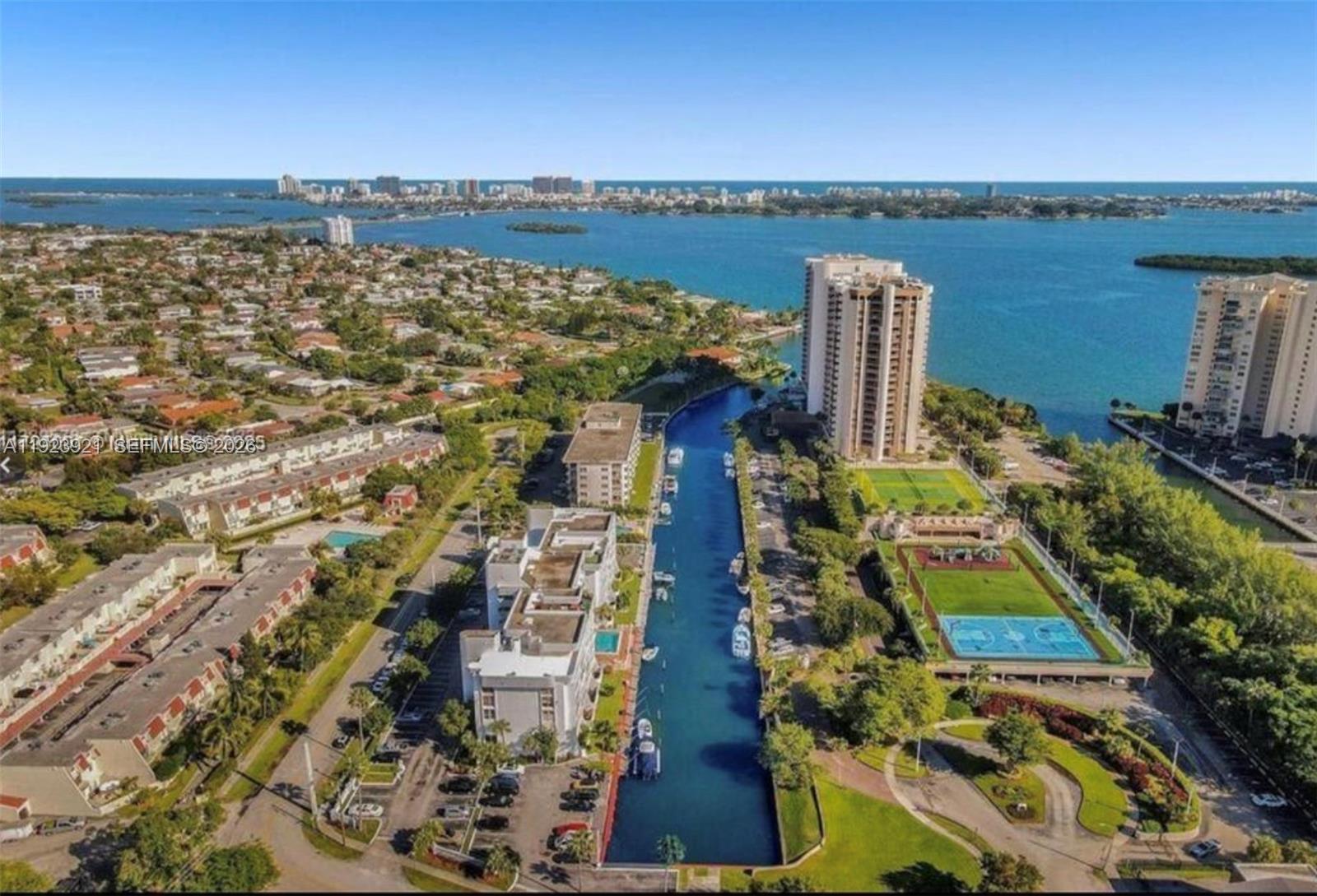 An aerial view shows a canal between residential buildings, tennis courts, and a distant bay — 1650 NE 115th St # 405, Miami FL 33181, MIAMI, FL
