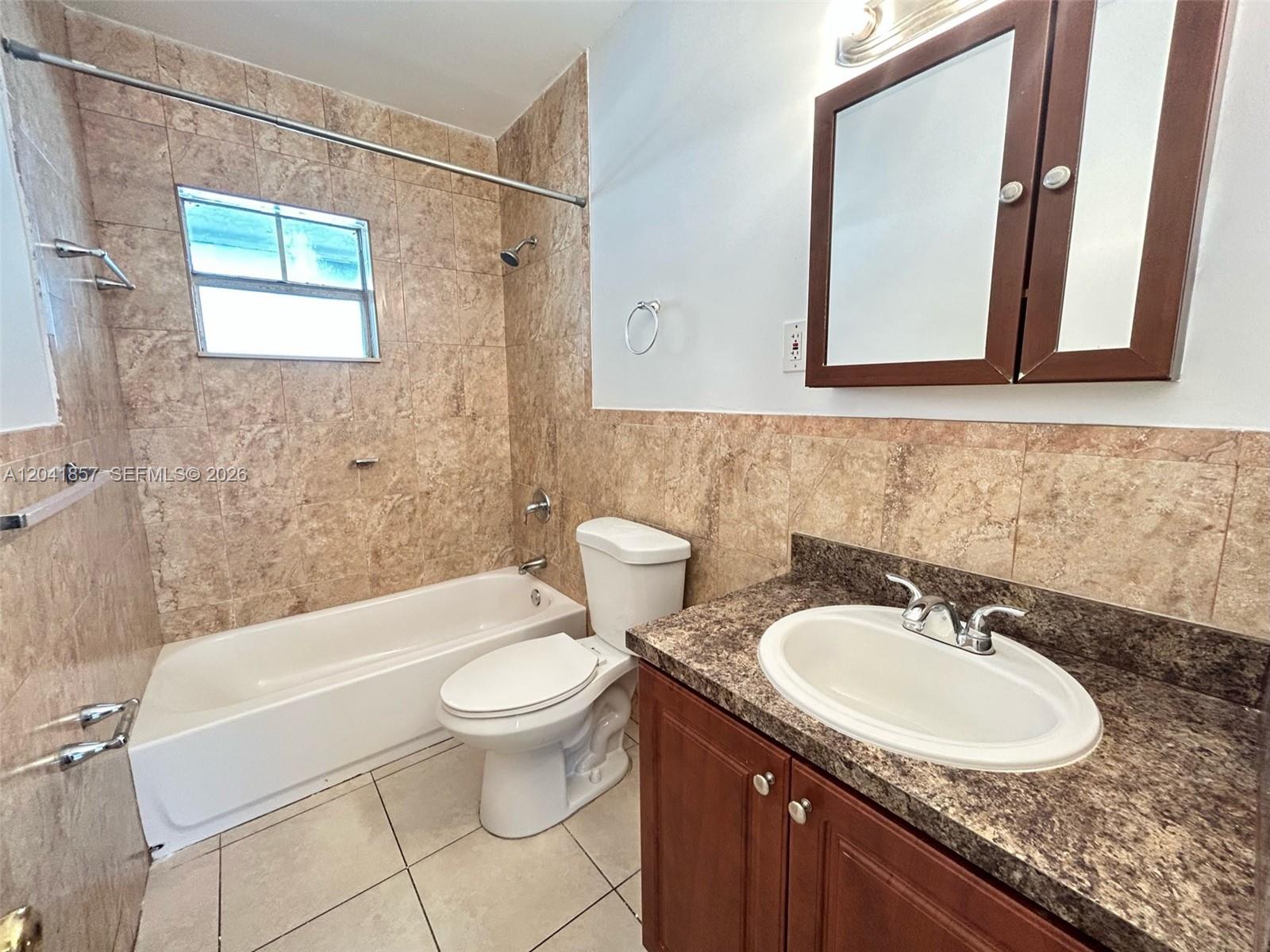 A bathroom features a tub-shower combo with tan tile, a toilet, and a wood vanity — 990 Phippen Waiters Rd, Dania Beach FL 33004, DANIA BEACH, FL