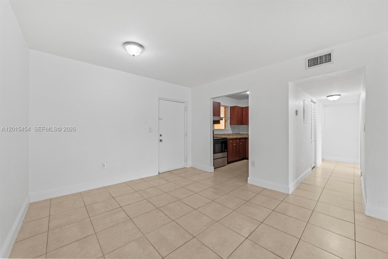 A white-walled room features beige tile flooring, a doorway to a kitchen, and a hallway — 525 W 69th St # 306, Hialeah FL 33014, HIALEAH, FL