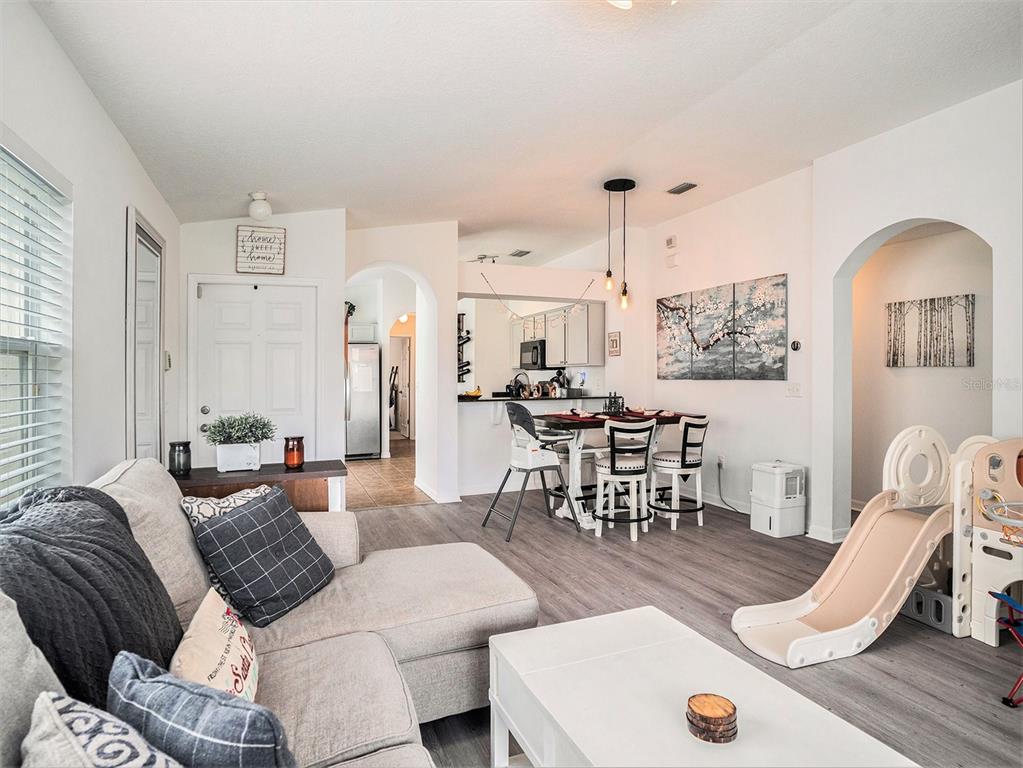 A living room and dining area with light wood-look flooring, white walls, and a kitchen — 7833 CARRIAGE POINTE DRIVE, GIBSONTON, FL