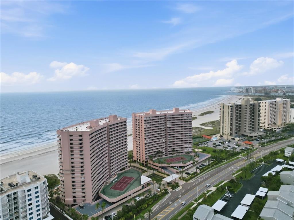 An aerial view of two tall pink beachfront condominium buildings with tennis courts and parking — 1310 GULF BOULEVARD UNIT 18C, CLEARWATER, FL