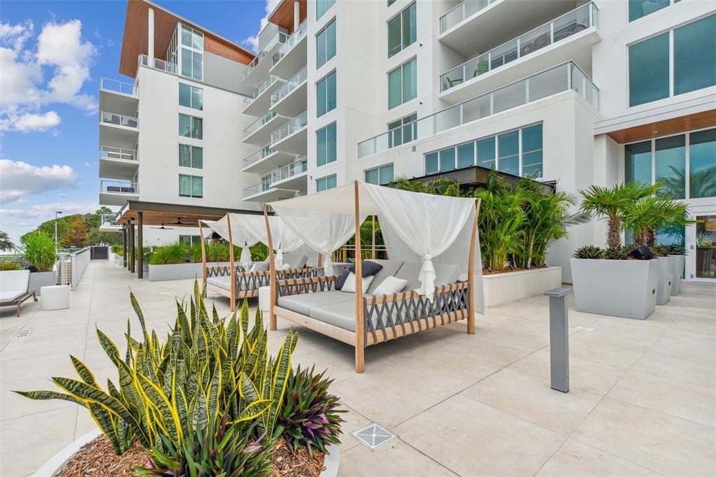 A modern outdoor patio features several shaded daybeds, potted plants, and a multi-story building exterior — 1020 SUNSET POINT ROAD UNIT 310, CLEARWATER, FL